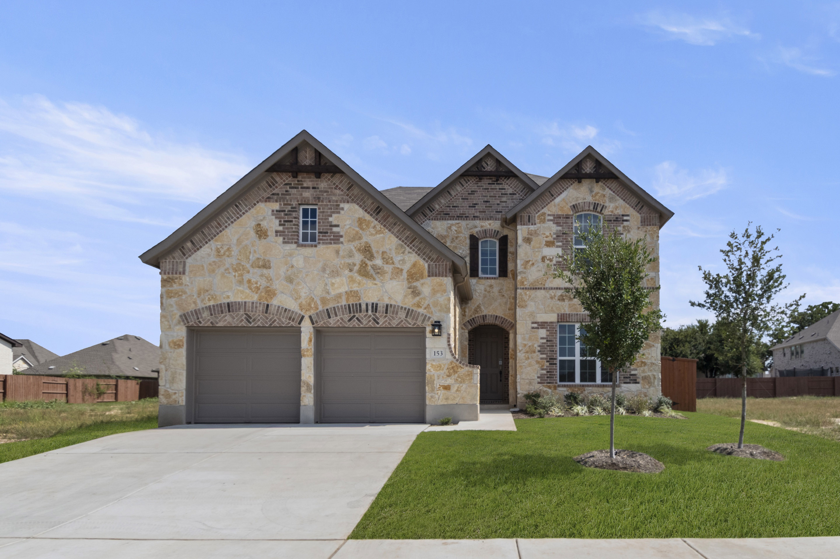 Front exterior of two-story brick house with double brown garage doors, green landscape and a tree.
