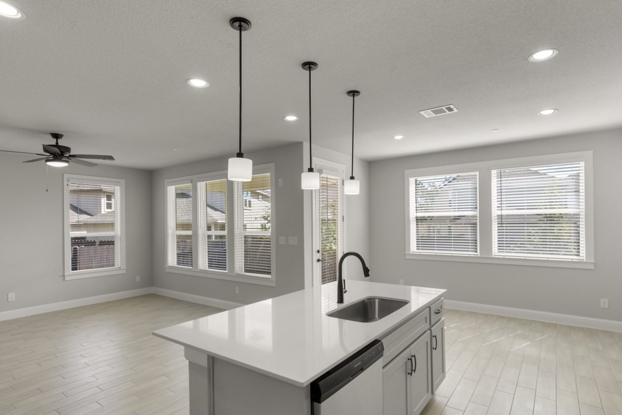 Kitchen and living room interior with white countertop kitchen island and tan vinyl flooring, and white-framed windows.