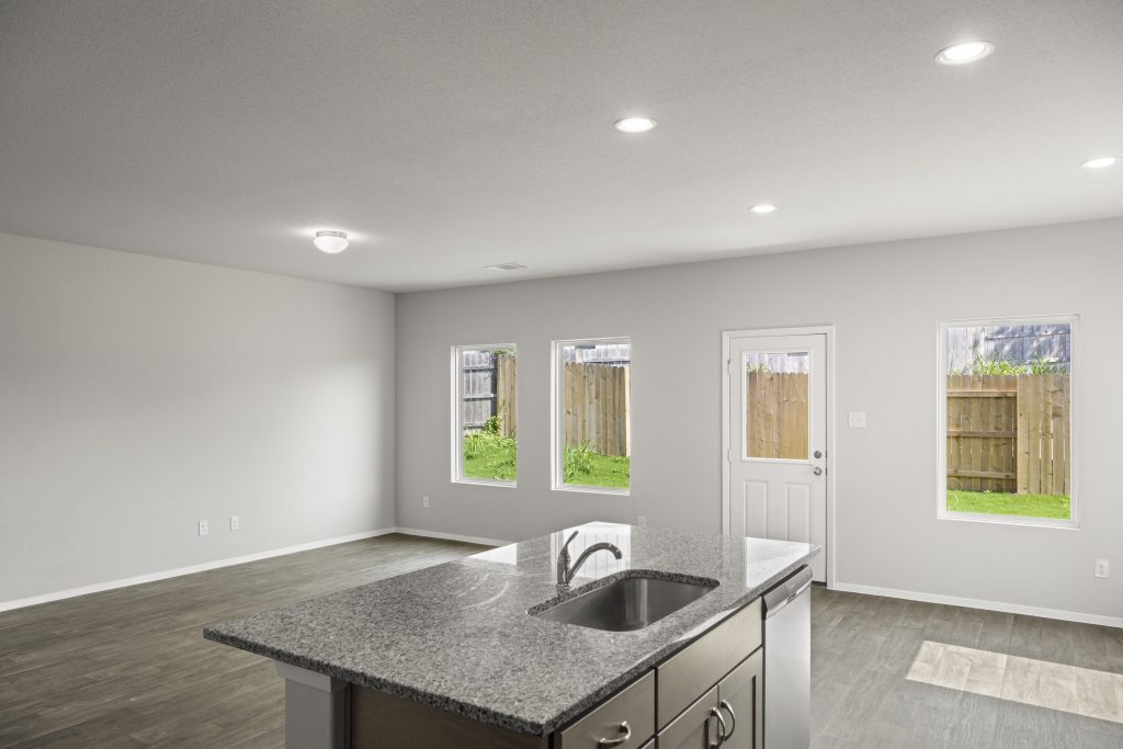 A kitchen and living room with a grey marble countertop island, vinyl flooring, white painted walls, white-framed window, and a white door.