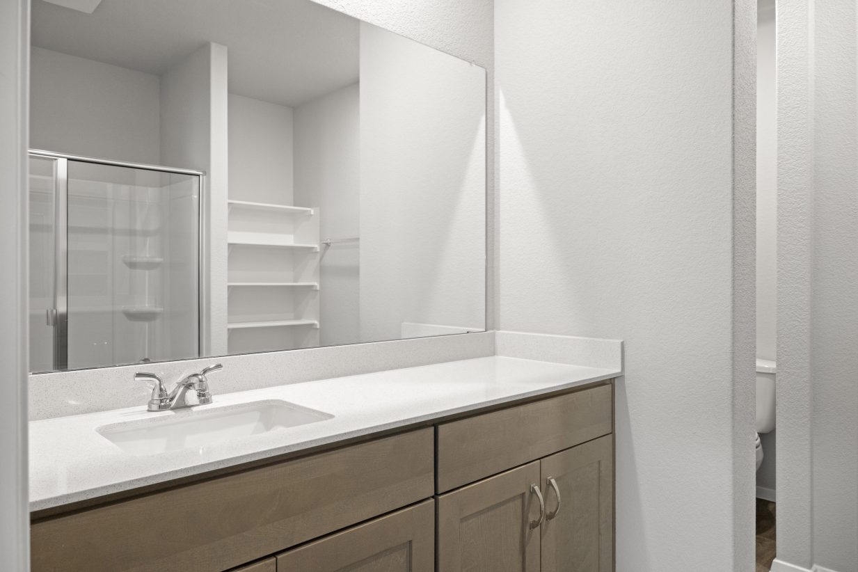 A bathroom with a white countertop, mirror, and brown cabinets, and white painted walls.