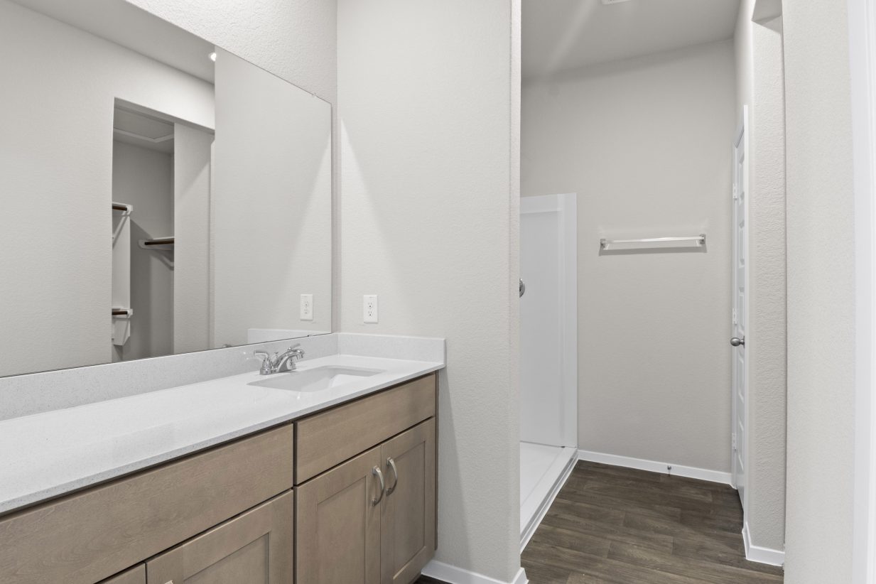 Interior image of a bathroom with a vanity with white countertops and brown cabinets, white walls, a mirror, wood-like vinyl flooring.