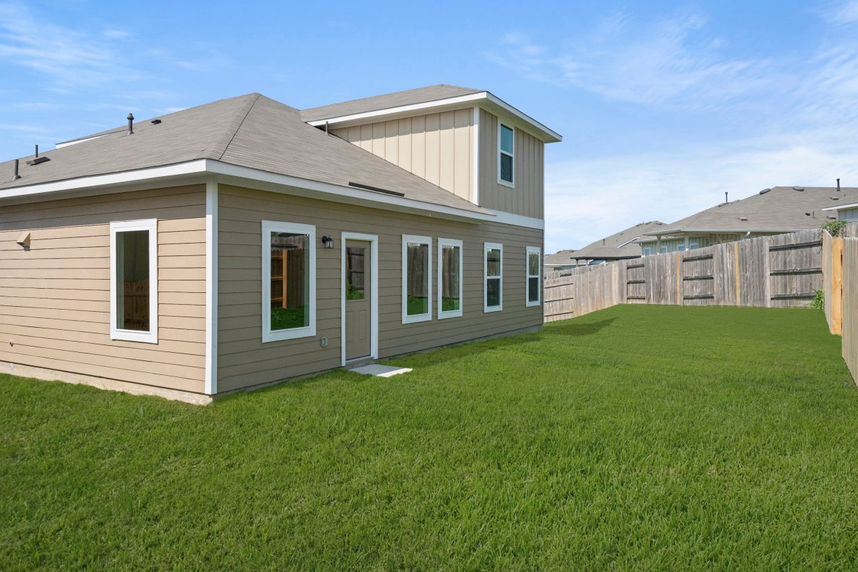 Back exterior of tan two-story home with white framing, green grass and brown fence.
