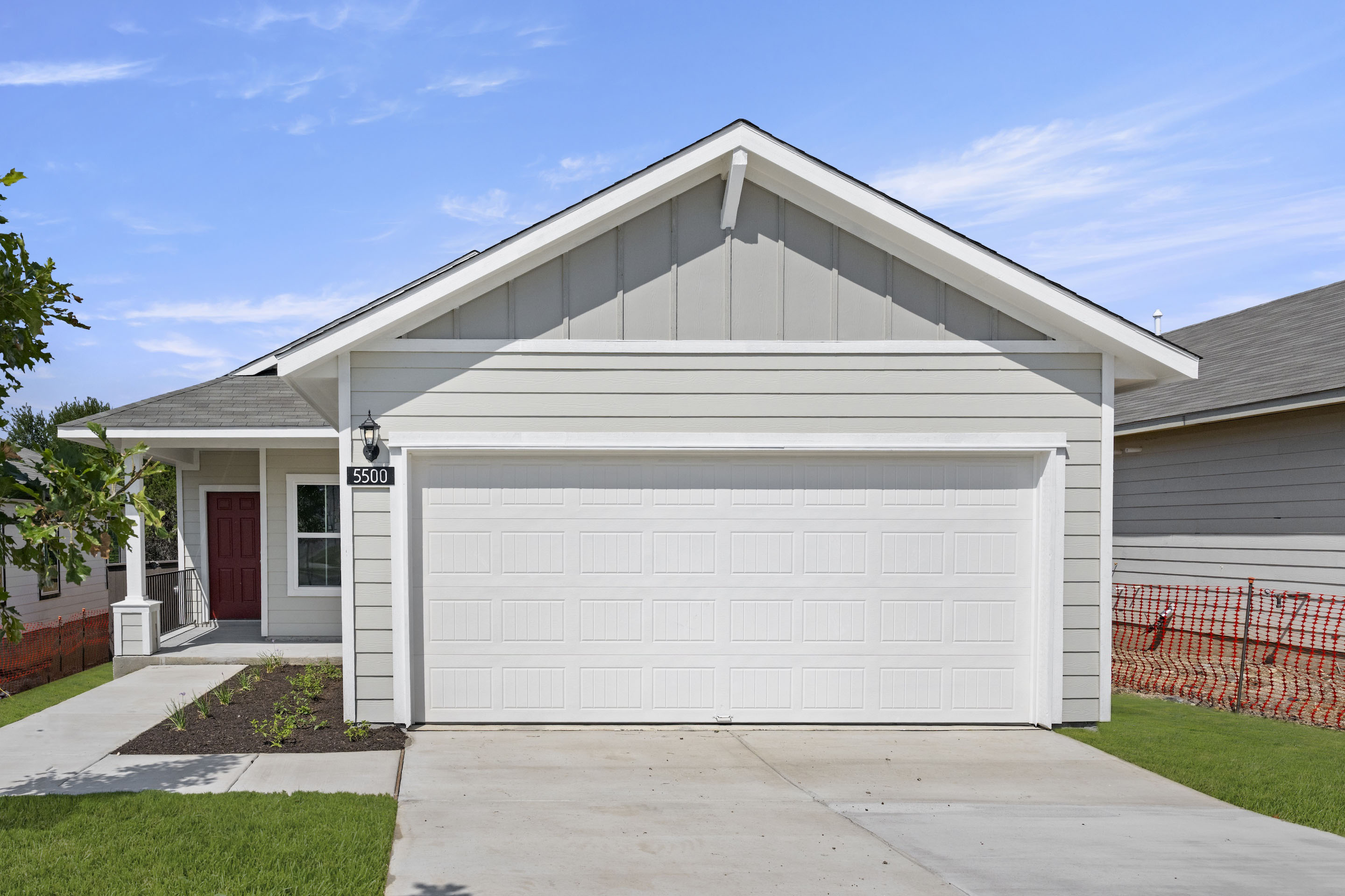 Front exterior of gray single-story home with red front door, a white garage and green landscape.