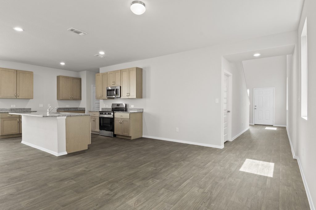 Interior image of kitchen with tan cabinets and granite countertops and living room with vinyl flooring.