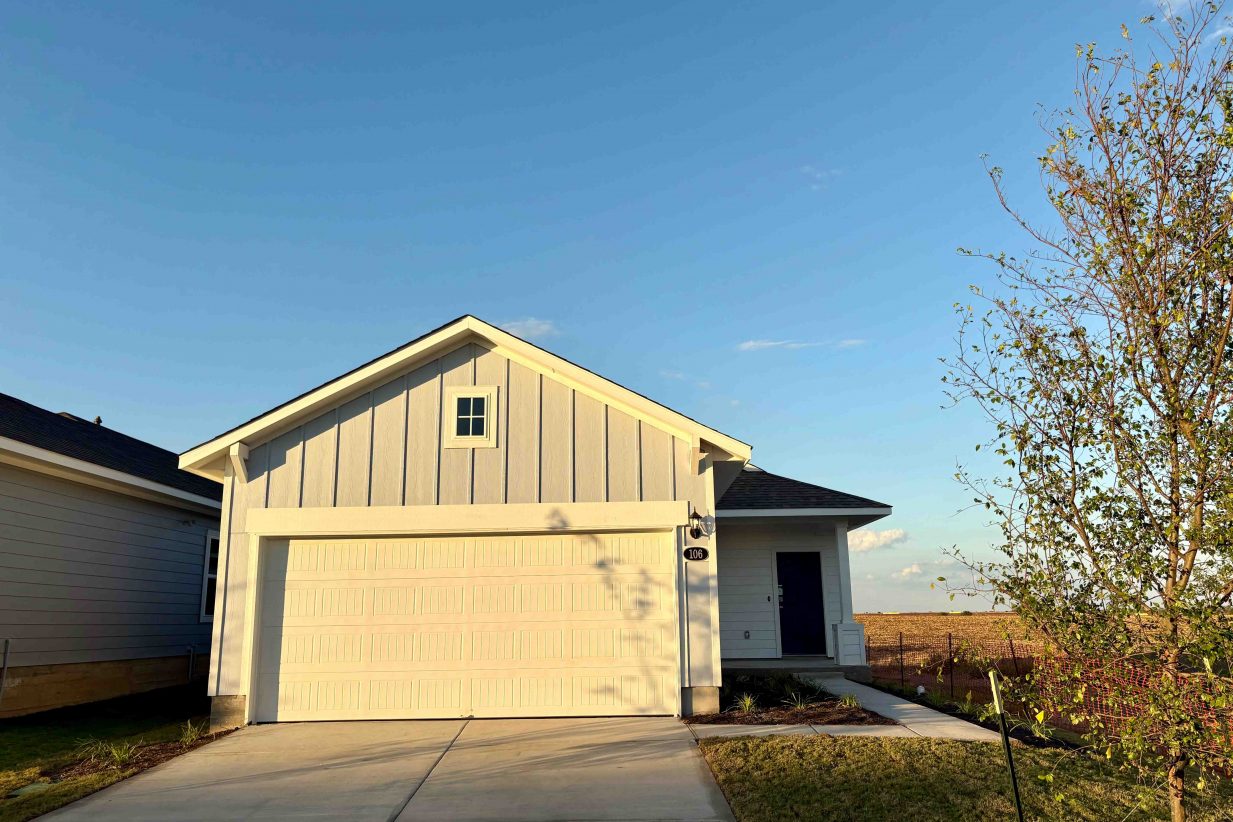 Image of a front of blue single-story home with white garage and driveway with a dirt landscape. and blue sky in the background.