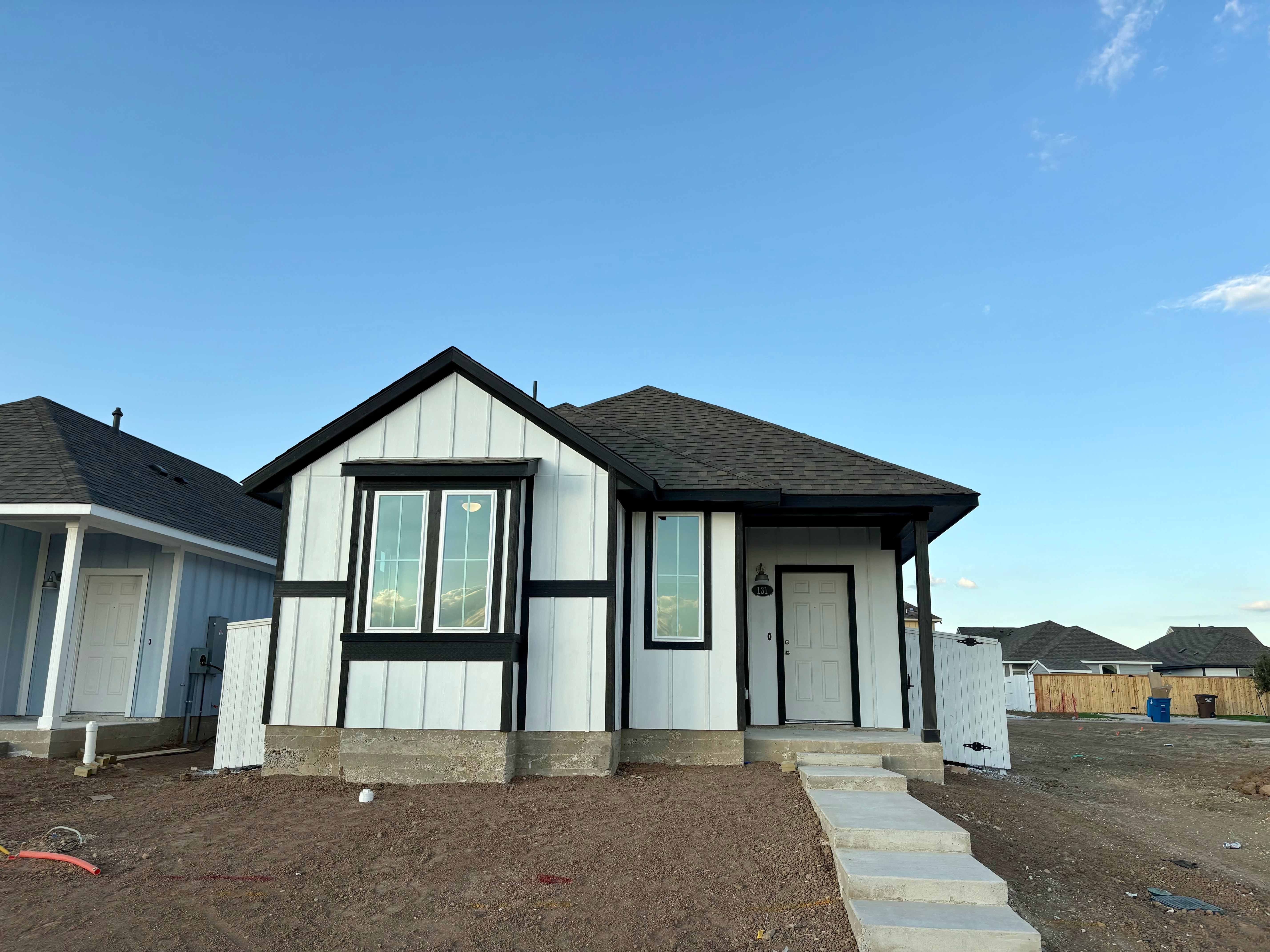 Image of front of a white farmhouse-style single story home with black trimming on a dirt lot and blue sky in the background.