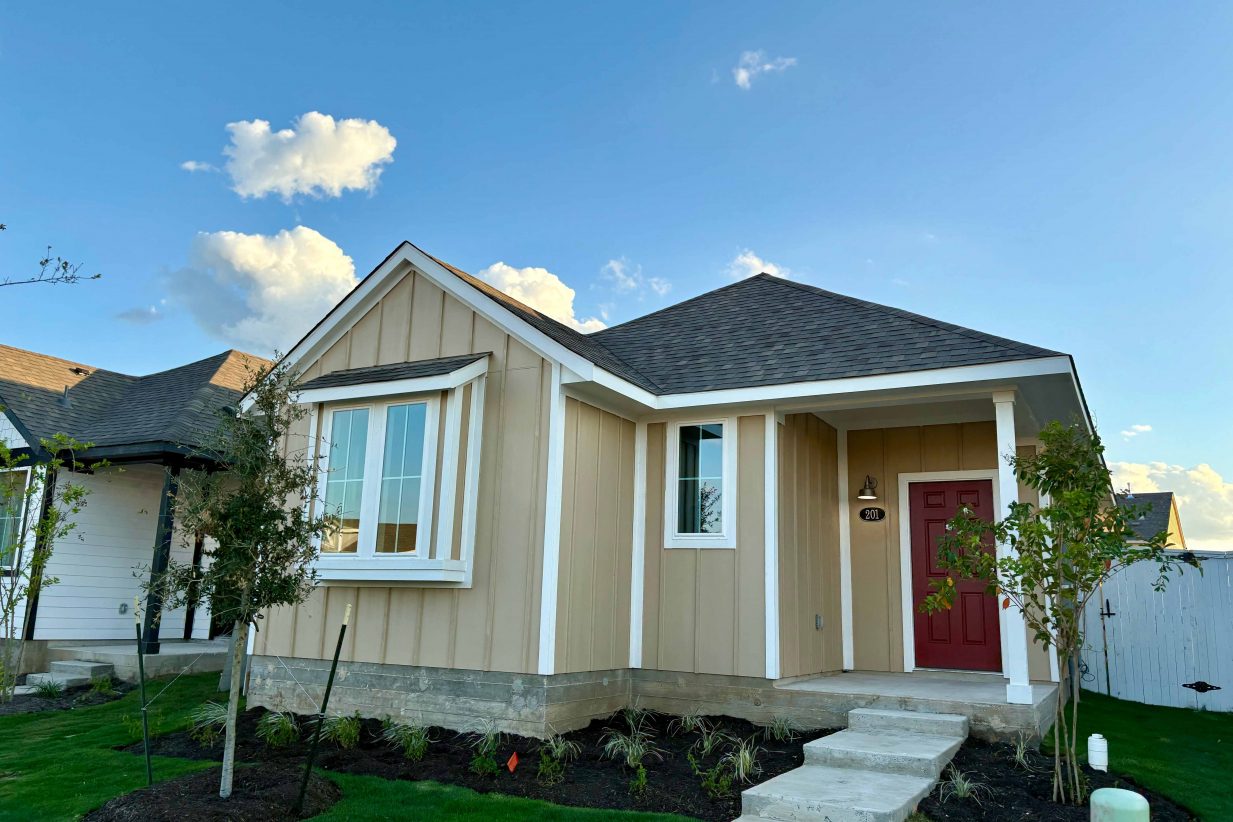 Image of front of yellow single-story house with a red front door, walk way, and green landscape with. a blue sky in the background.