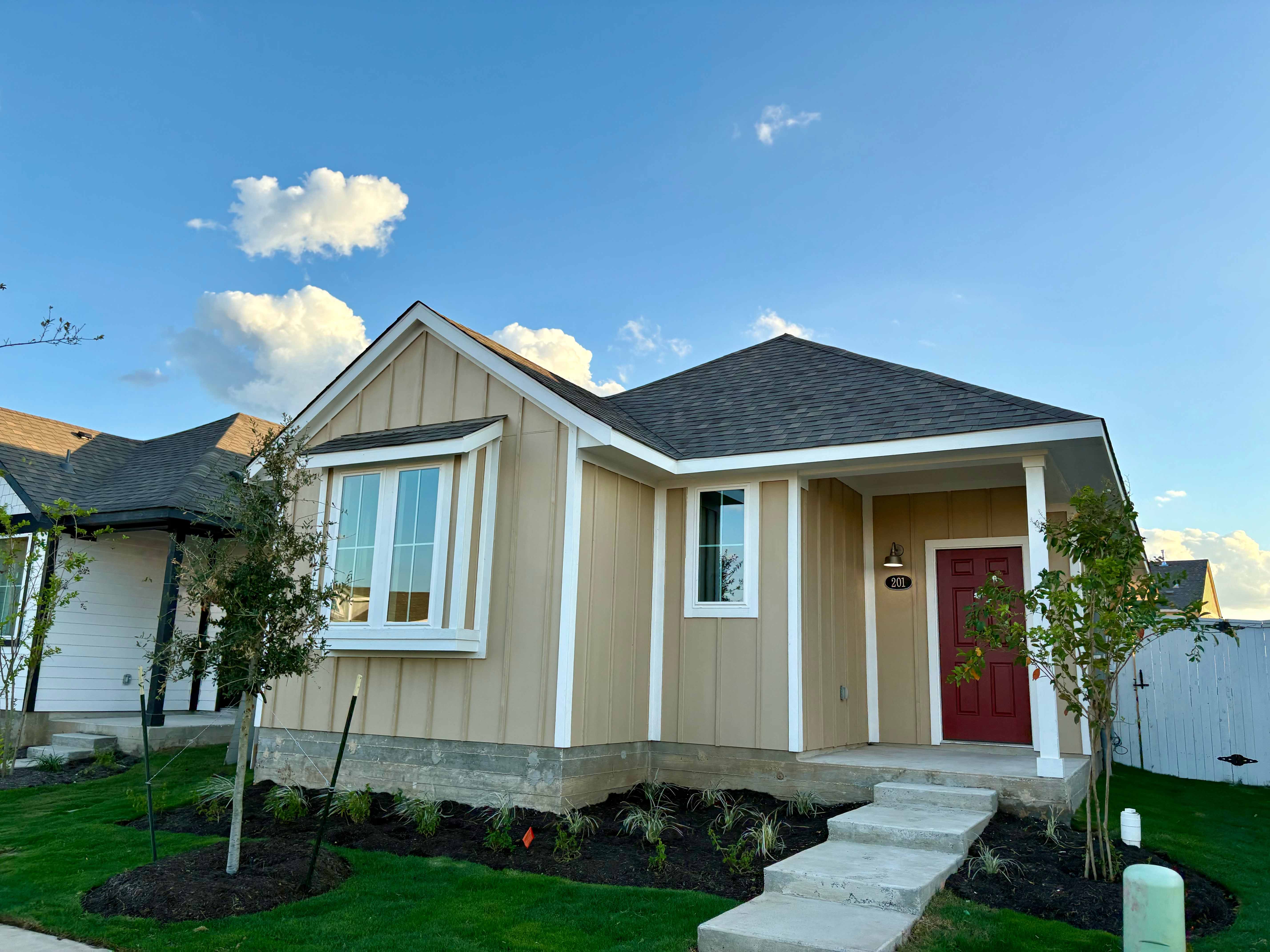 Image of front of yellow single-story house with a red front door, walk way, and green landscape with. a blue sky in the background.
