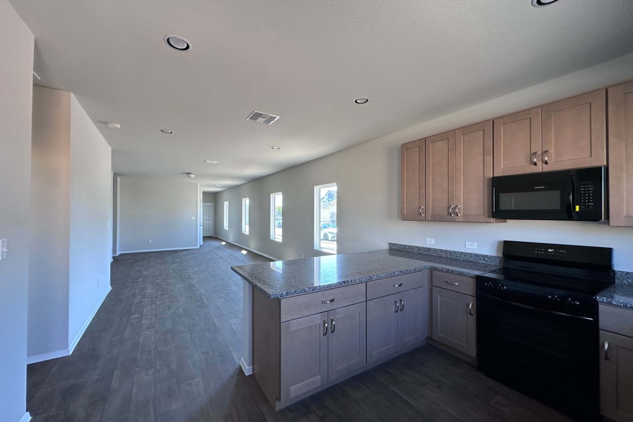An image of a kitchen with gray and white speckled countertop, grey cabinets, black appliances looking into the living room with vinyl wood-like flooring.