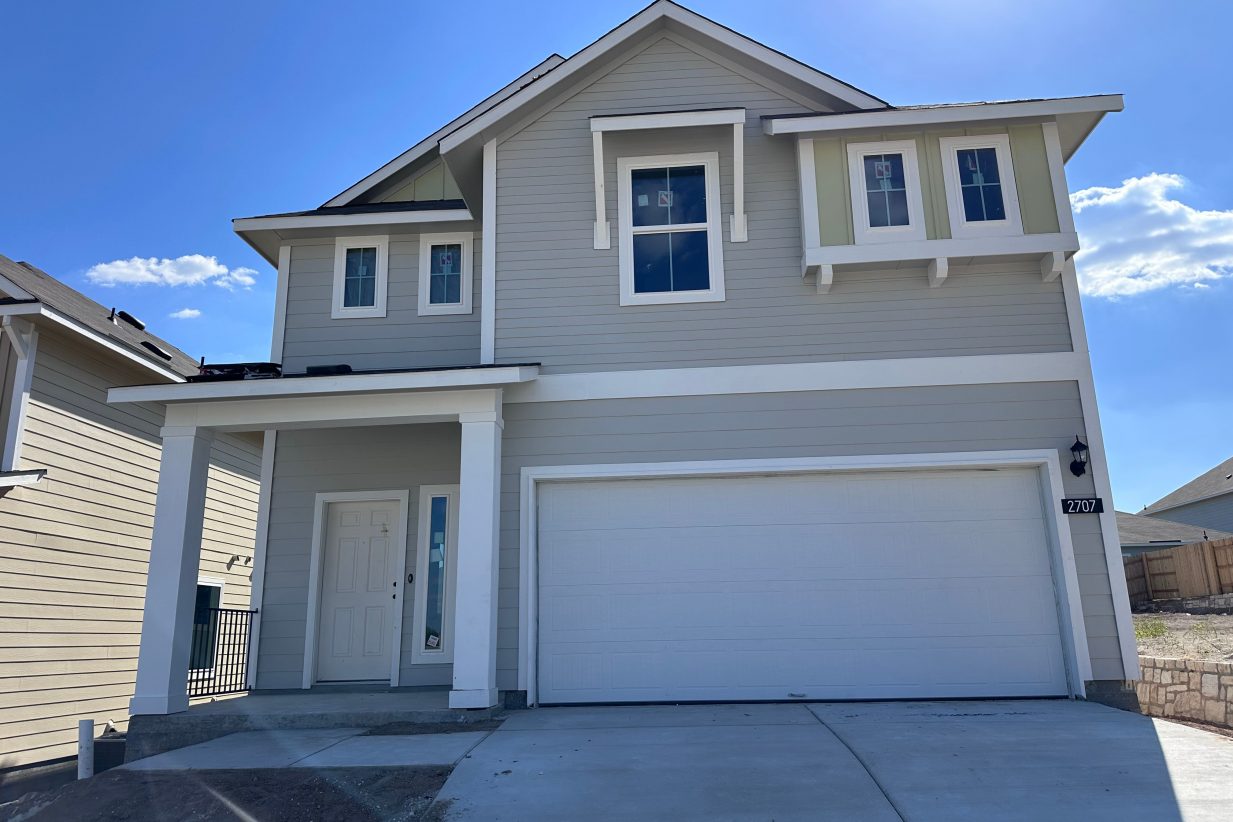An image of grey and yellow two-story home with a white garage, driveway, dirt landscape, and blue sky in the background.