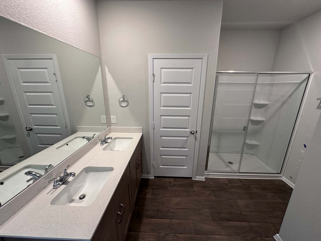 An image of a bathroom with white countertops, brown cabinets, a glass shower, white painted walls, and vinyl wood-like vinyl flooring.
