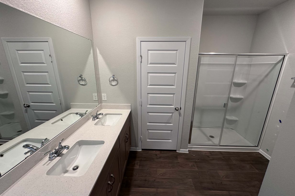 An image of a bathroom with white countertops, brown cabinets, a glass shower, white painted walls, and vinyl wood-like vinyl flooring.