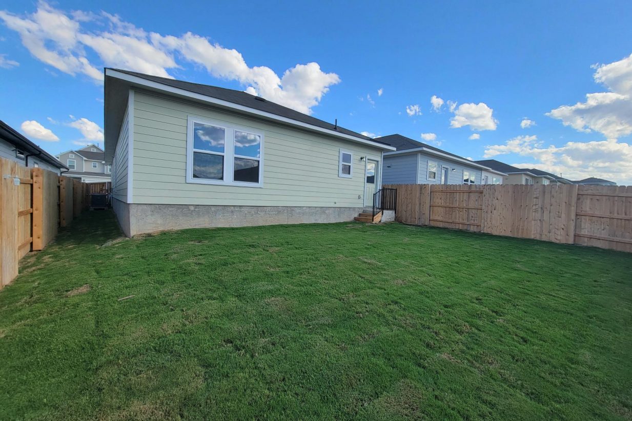 Image of exterior of one story yellow home, green grass, wood fence, and sky