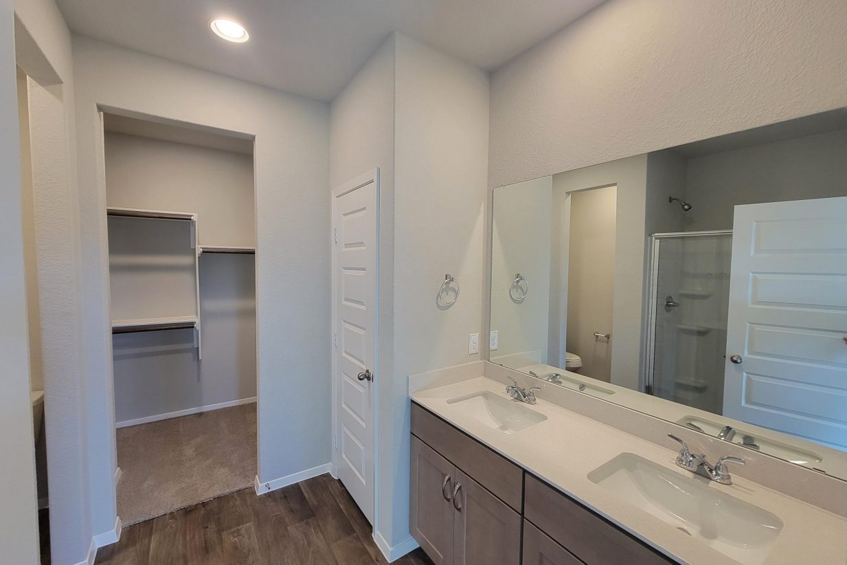 Image of home primary bathroom with dual vanity with white countertop and light brown cabinets, mirrow, white walls, wood-look floor, and doorway to walk-in closet