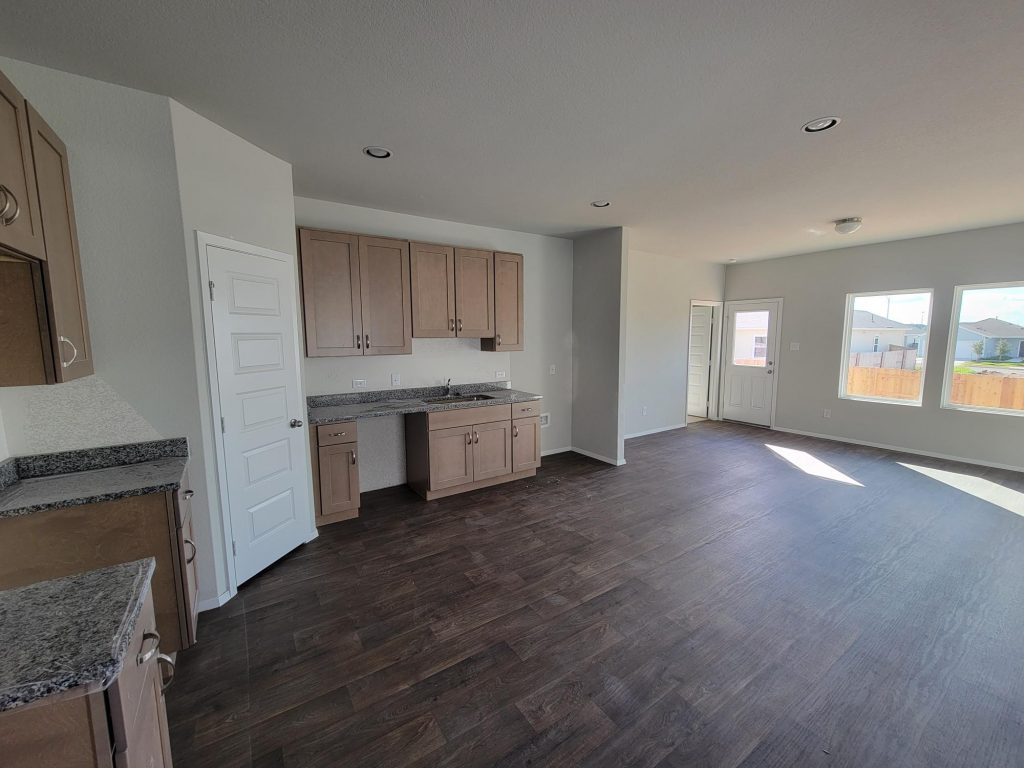 Image of home living area and kitchen with light brown cabinets, wood look floor, white walls, and windows