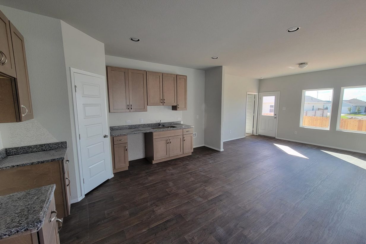 Image of home living area and kitchen with light brown cabinets, wood look floor, white walls, and windows