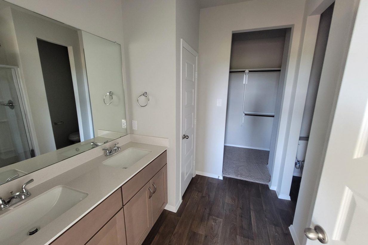 Image of home bathroom with double vanity with light brown cabinets and white countertops, mirror, and doorway to walk-in closet