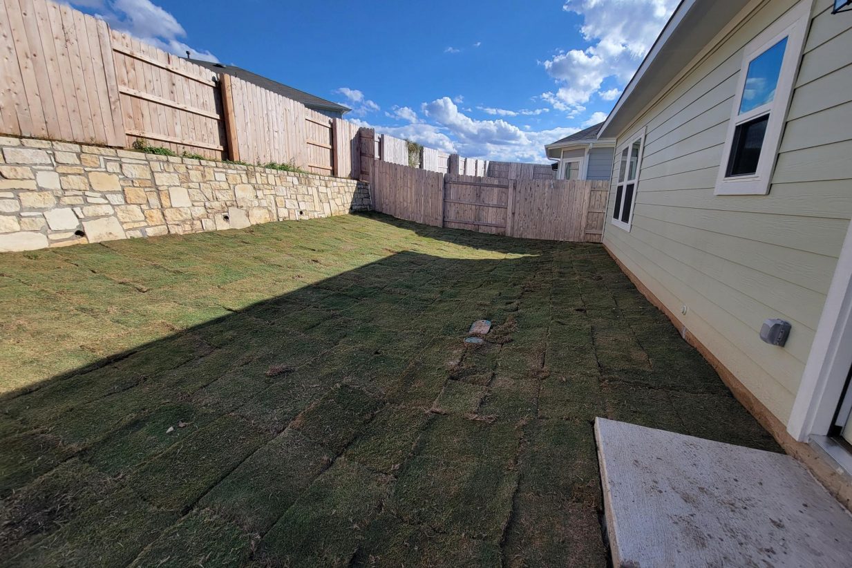 Image of home exterior with retaining wall on one side and one story home with green grass, wood fence, and sky