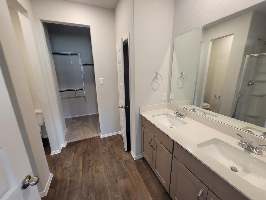 Image of home bathroom with dual vanity with white countertop and light brown cabinet, mirror, wood-look floor, and doorway to walk-in closet