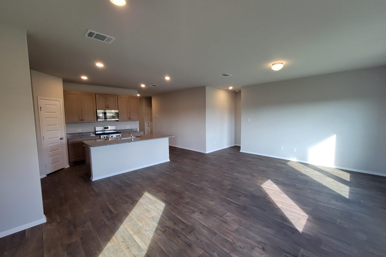 Image of home living area and kitchen with center island and brown cabinets. Wood-look floor and white walls