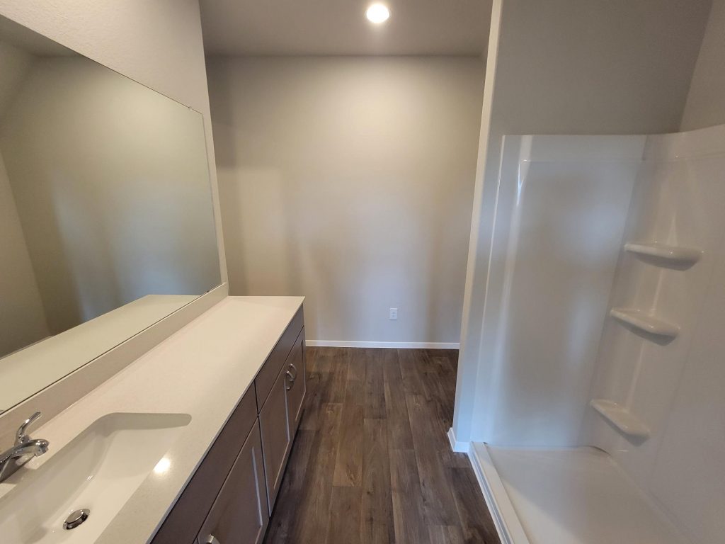 Image of home bathroom with vanity with white countertop and brown cabinets, walk-in shower, and wood-look floor