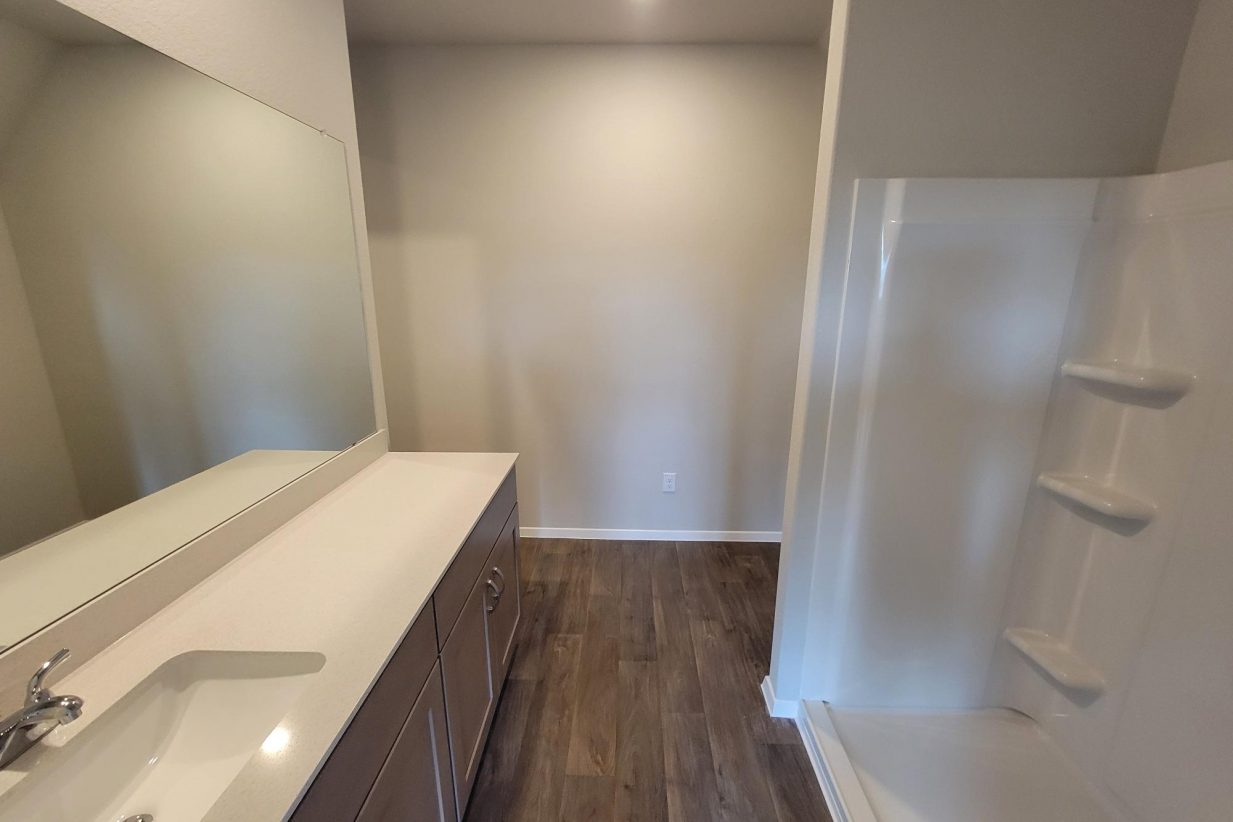 Image of home bathroom with vanity with white countertop and brown cabinets, walk-in shower, and wood-look floor