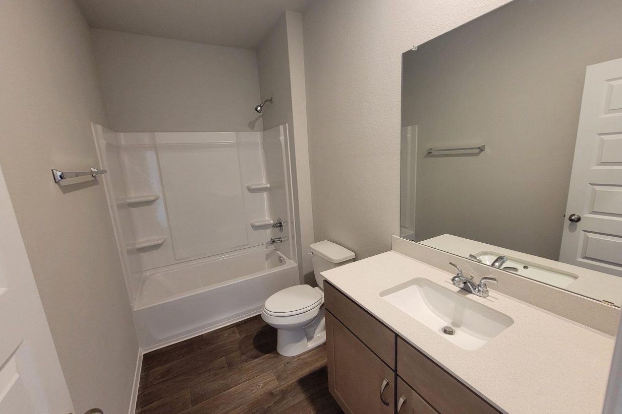 Image of home bathroom with vanity with white countertop and light brown cabinet, mirror, wood look floor, toilet, and tub