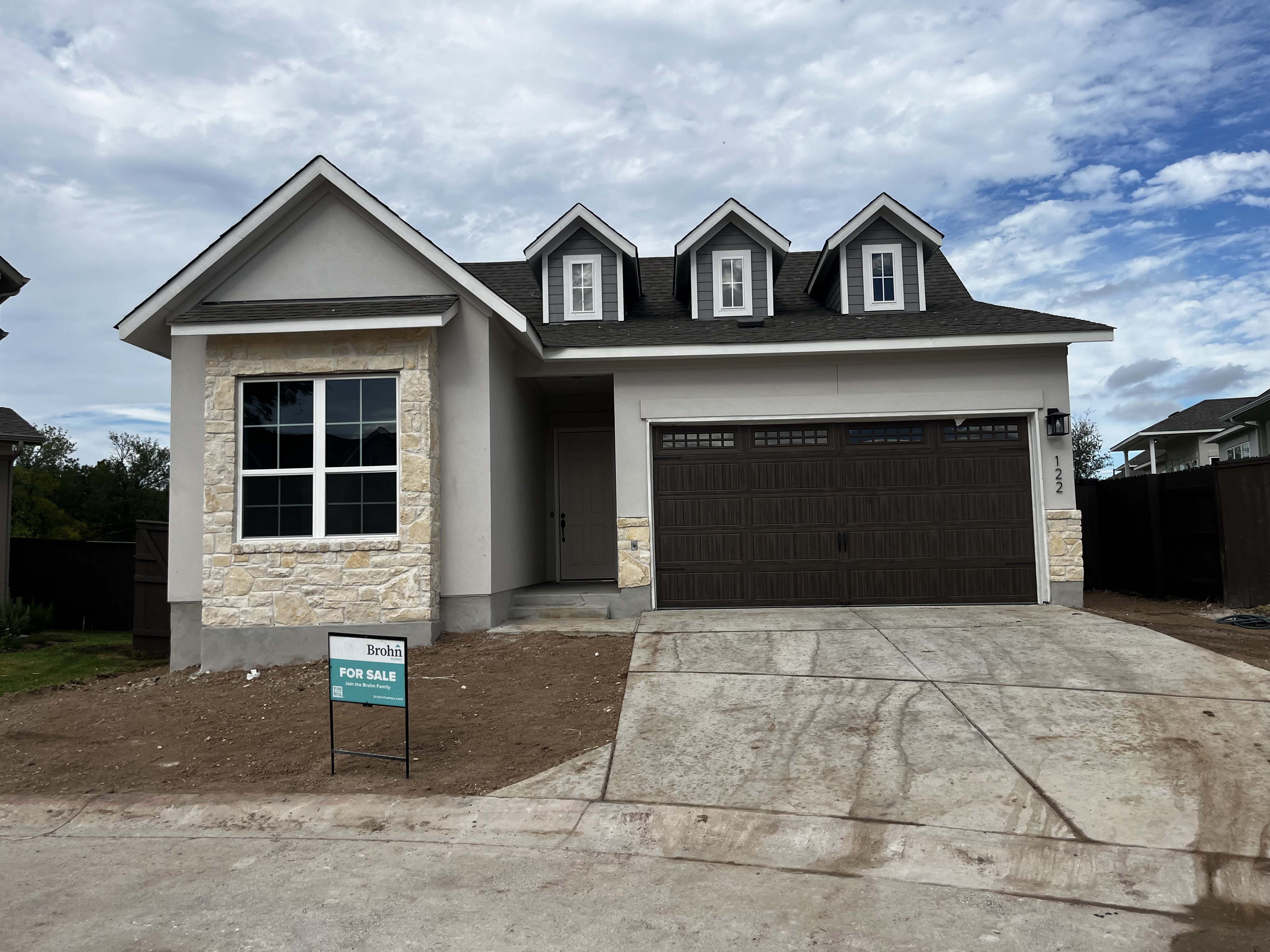 Front exterior of gray and brick accented single-story house with a brown two-car garage and dirt landscape.