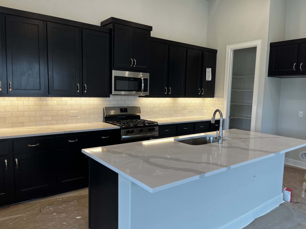 A kitchen with white marbled countertops, brown cabinets, steel appliances, white blacksplash tiles, and a kitchen pantry.