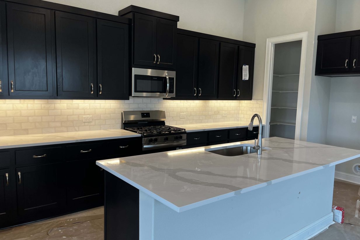 A kitchen with white marbled countertops, brown cabinets, steel appliances, white blacksplash tiles, and a kitchen pantry.