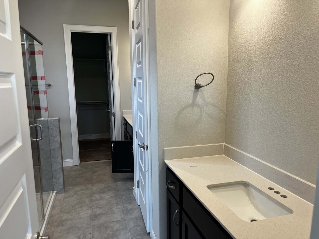 A bathroom with gray floor tiles, a glass shower, a vanity with white countertop and brown cabinets and gray painted walls.