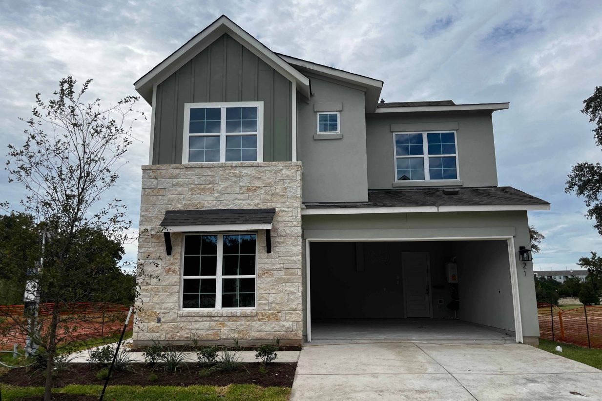 Image of front of a green two-story house with brick accents, open garage door, and green landscape.