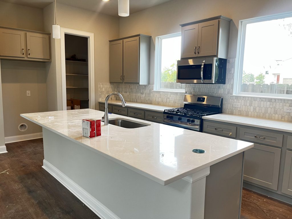An image of a kitchen with brown vinyl flooring, white and grey backsplash tiling, grey cabinets, and white countertops, a central island with white countertop, and steel appliances.