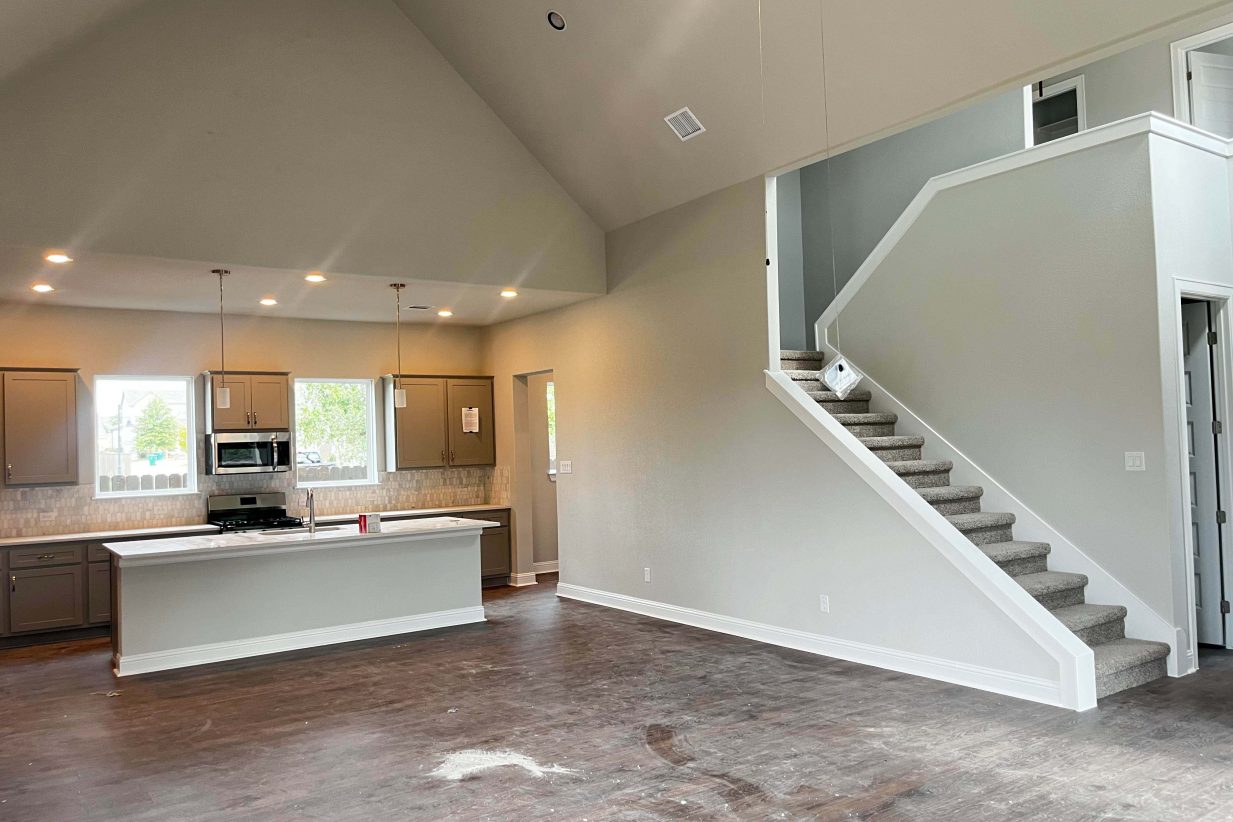 An image of a kitchen and living room with brown vinyl flooring, cream colored painted walls, white base boards, and carpet stairwell.